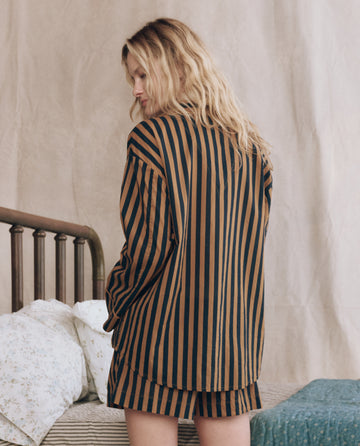 A person with long, wavy blonde hair stands by a bed, wearing THE GREAT. F25 SLEEP's The Pajama Shirt in Club Stripe: a vintage-inspired brown and black vertically striped pajama shirt and shorts set.