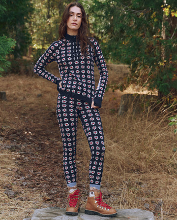 A woman stands outdoors among trees and dry grass wearing THE GREAT. F25 TGO's The Merino Legging in Frosted Daisy—slim, floral leggings paired with brown hiking boots, perfect for a slopeside adventure.