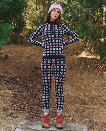 Standing outside on a tree stump, this person wears THE GREAT. F25 TGO's The Merino Quarter Zip in Frosted Daisy—paired with patterned thermals, red hiking boots, and a knit hat, the perfect slopeside companion among dry grass and trees.