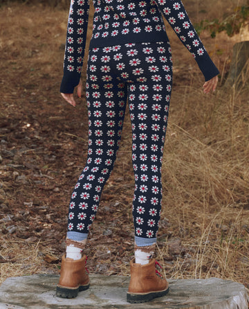 Standing outdoors on a rock, this person wears THE GREAT. F25 TGO's The Merino Legging in Frosted Daisy—a slim, slopeside-ready legging featuring a white and red floral pattern. Brown boots complete the look amid dry grass and leaves.