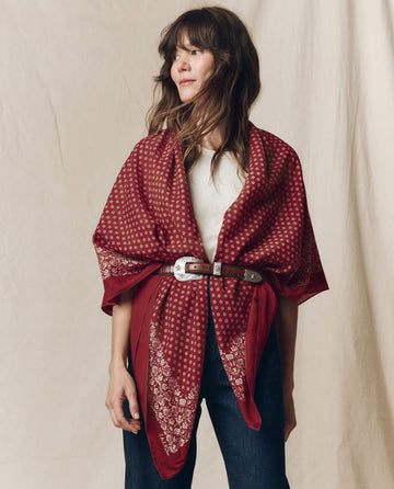 A woman wearing THE GREAT. F26 BIG BANDANA in maroon, styled as a shawl belted at the waist over a white top and jeans, standing in front of a beige backdrop.