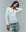 A person with long braided hair in THE GREAT. F25 BABY RIB "The Baby Rib Henley" in True White and light blue jeans poses against a plain light gray background, tilting their head and touching their neck.