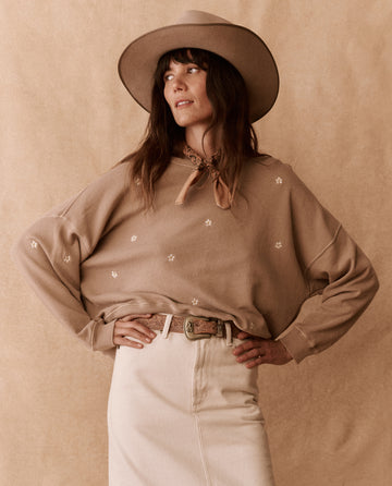 A woman in THE GREAT. PF25 EMB KNITS’ The Slouch Sweatshirt—Khaki with Daisy Embroidery—stands hands-on-hips, paired with a cream skirt and wide-brim hat, against a coordinating tan backdrop.