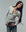 A person with long braided hair, wearing THE GREAT. F25 BABY RIB The Baby Rib Henley in Heather Grey and blue jeans, stands against a plain background, smiling and tilting to the side.
