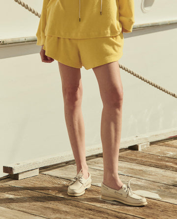 Person standing on a wooden dock, wearing THE GREAT. SU26 The Micro Terry Swing Short in Margarita, paired with a matching yellow top and beige boat shoes. Upper body is out of frame.