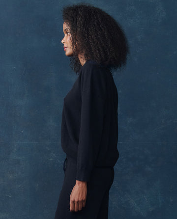 A woman with curly hair stands in profile against a dark blue textured background, wearing THE GREAT. H25 CASHMERE’s The Cashmere Crew in Navy.