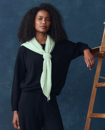 A woman stands by a wooden ladder in black attire, wearing THE GREAT. H25 CASHMERE’s The Cashmere Kerchief Scarf in Mint, tied around her neck, against a dark blue background.