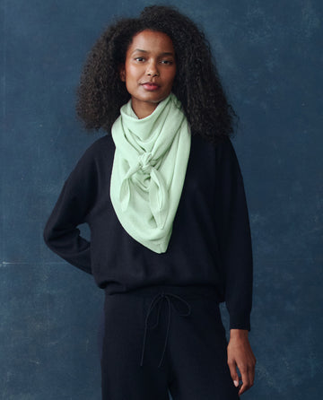 A woman with curly hair wears a black long-sleeve outfit and The Cashmere Kerchief Scarf in Mint by THE GREAT. H25 CASHMERE, tied in front, against a dark blue background.