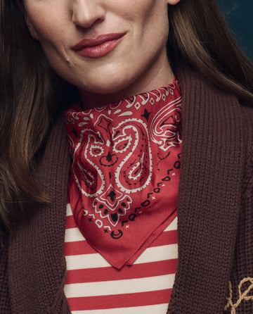 A woman wearing THE GREAT. F25 COLLECTORS The Silk Bandana in True Red around her neck, paired with a red and white striped shirt and a brown knitted cardigan.