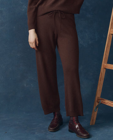 Wearing THE GREAT. H25 CASHMERE’s The Cashmere Lantern Pant in Chocolate, this look exudes refined luxury loungewear as they stand by a wooden ladder against a blue textured backdrop.