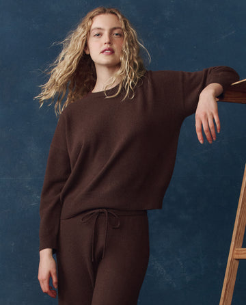 A person with long, wavy blond hair wears THE GREAT. H25 CASHMERE "The Cashmere Crew" in Chocolate and matching pants, standing by a wooden ladder against a dark blue background.