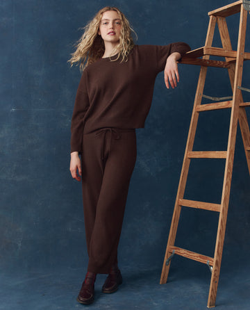 Wearing THE GREAT. H25 CASHMERE’s The Cashmere Lantern Pant in Chocolate, a person stands with one arm on a wooden ladder against a blue background, pairing the luxury pant with a brown sweater for an elevated loungewear look.