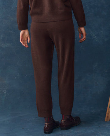A person faces away from the camera, wearing THE GREAT. H25 CASHMERE's The Cashmere Lantern Pant in Chocolate with a matching top, dark patterned socks, and brown loafers, set against a blue textured background.