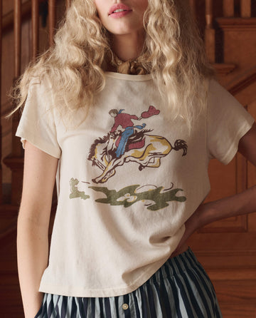 Someone with long wavy hair wears THE GREAT.'s The Little Tee W/ Rider Graphic in washed white, featuring a bucking horse print, paired with a blue and white striped skirt.