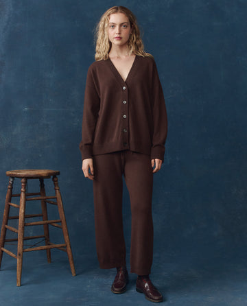 A woman stands indoors against a dark blue background, wearing THE GREAT. H25 CASHMERE The Cashmere Slouch Cardigan in Chocolate with matching pants and burgundy shoes, next to a wooden stool.