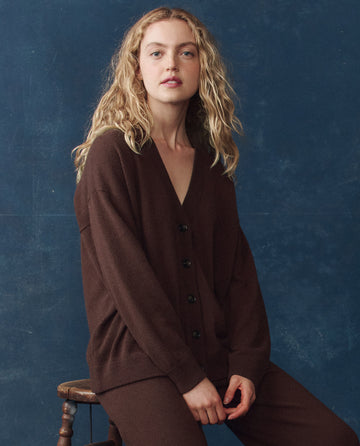 A woman with wavy blonde hair wears THE GREAT. H25 CASHMERE’s The Cashmere Slouch Cardigan in Chocolate while sitting on a wooden stool against a blue background.