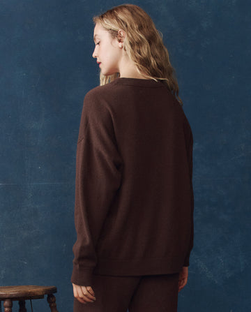 A person with wavy blonde hair wears THE GREAT. H25 CASHMERE’s The Cashmere Slouch Cardigan in Chocolate, standing with their back to the camera against a blue background beside a small wooden stool.