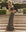 A woman stands on stone steps outdoors, surrounded by greenery and a fountain, wearing THE GREAT. SP25 SWIM’s The Pareo. -- Leopard wrap skirt and a strapless black top.