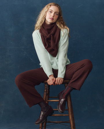 A woman with blonde curly hair sits on a wooden stool, wearing a light mint sweater, brown pants, brown loafers, and THE GREAT. H25 CASHMERE The Cashmere Kerchief Scarf in Chocolate against a dark blue background.