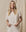 A person with long blonde hair smiles while wearing The Boxy Crew in Washed White by THE GREAT. CORE KNITS, against a beige fabric backdrop.