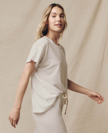 Wearing THE GREAT. CORE KNITS' The Boxy Crew in Washed White, a 100% cotton top, the person poses against a beige backdrop, flaunting this vintage tee ideal for any casual occasion.