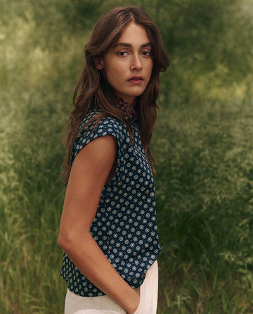 In a grassy outdoor setting, a person with long hair wears The Boxy Trail Tank - Micro Daisy by THE GREAT. SP25 TGO and white pants while looking toward the camera.