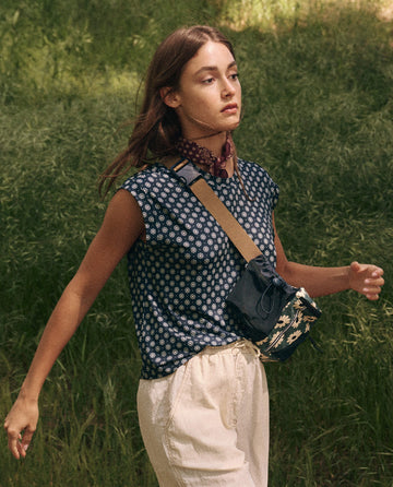 A woman strolls along the path, her Boxy Trail Tank by THE GREAT. SP25 TGO under a patterned top and light pants. With a small crossbody bag and loose hair flowing around her scarf-draped neck, she embodies thoughtful elegance in outdoor apparel.