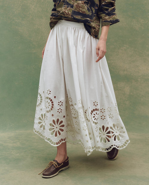 Someone stands against a green backdrop in THE GREAT. SU25 The Super Bloom Eyelet Skirt—White, styled with a printed top and brown boat shoes for a fresh, Super Bloom-inspired look.