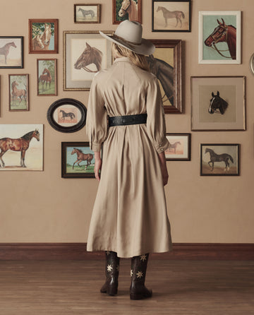 Wearing a beige dress, THE GREAT. STETSON SP26 The Gus 6X Hat in Ranch Tan, and cowboy boots, a person stands with their back to the camera facing a wall adorned with framed horse pictures.