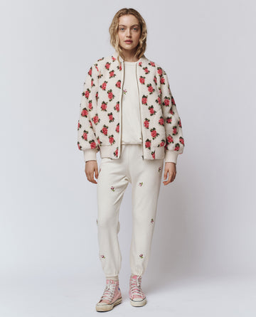 A person stands against a white background wearing THE GREAT. H25 ROSEBUD’s Zip Front Plush Bomber in Cream Rose, featuring an embroidered rose motif, paired with joggers and light pink high-top sneakers.