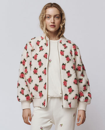 A woman wears THE GREAT. H25 ROSEBUD “The Zip Front Plush Bomber – Cream Rose” with matching pants and a white tee, all featuring pink rose prints and an embroidered motif, standing against a plain background.