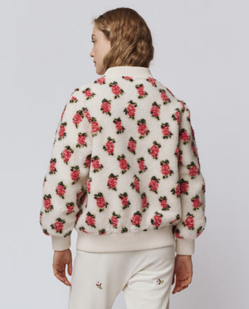 A person with light brown hair models THE GREAT. H25 ROSEBUD's "The Zip Front Plush Bomber -- Cream Rose," featuring a pink rose motif, paired with matching cream pants and shown from the back against a plain background.