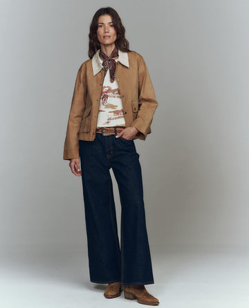 A person faces forward against a plain backdrop, wearing THE GREAT. F25 D1 The Waxed Swingy Jacket in Walnut with a corduroy collar, wide-leg dark jeans, brown boots, a patterned shirt, and a scarf around their neck.