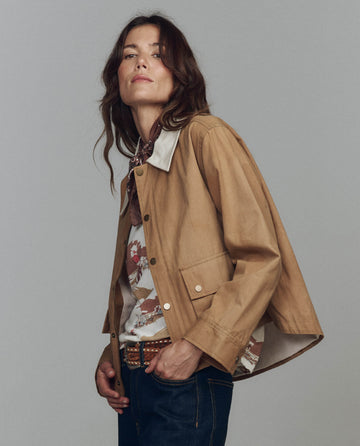 Wearing THE GREAT. F25 D1 The Waxed Swingy Jacket in Walnut over a patterned shirt, dark jeans, and a neck scarf, a person stands against a neutral gray background.