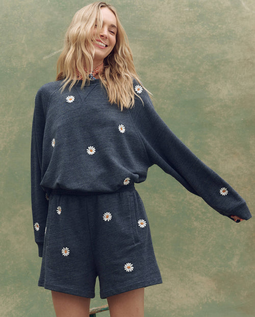 A person wearing THE GREAT. SU25 The Slouch Sweatshirt in heathered vintage blue with floating daisy embroidery stands smiling with one arm outstretched before a green background.