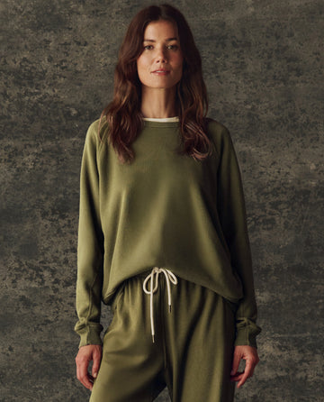 A woman faces forward in THE GREAT. H25 BRIGHT FLEECEBACK The Fleece Rookie Sweatshirt, Solid -- Army, paired with matching joggers, set against a textured gray background.