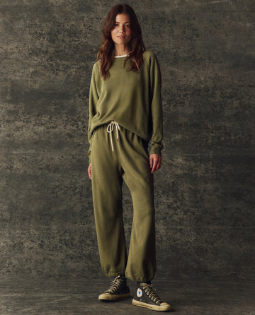 A person stands against a textured backdrop wearing THE GREAT. H25 BRIGHT FLEECEBACK The Fleece Sport Sweatpant in Army, paired with a coordinating sweatshirt and black high-top Converse sneakers.