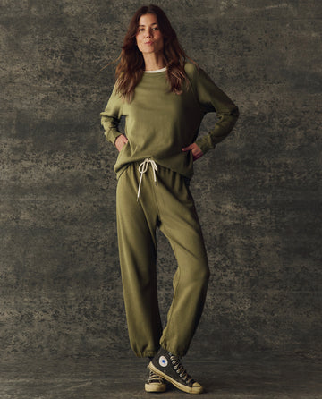 A woman stands against a textured backdrop wearing THE GREAT.'s The Fleece Rookie Sweatshirt in Army from the H25 BRIGHT FLEECEBACK collection, paired with sneakers and her hands in her pockets.