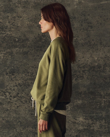 A woman with long brown hair stands in profile against a textured dark background, wearing THE GREAT. H25 BRIGHT FLEECEBACK’s The Fleece Rookie Sweatshirt in Solid Army and matching pants.