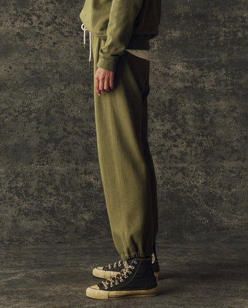 A person stands in profile wearing THE GREAT. H25 BRIGHT FLEECEBACK The Fleece Sport Sweatpant in Army, paired with a matching sweatshirt and black high-top sneakers against a textured dark background.
