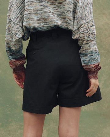 Person standing with their back to the camera, wearing THE GREAT. SU25 The Pleated Trouser Short in black and a multicolored long-sleeve sweater, set against a green mottled background.