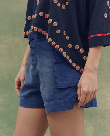 A person stands outdoors with their arm by their side, wearing THE GREAT. SU25 The Cargo Fatigue Short in Vintage Blue and a navy blue top with floral embroidery.