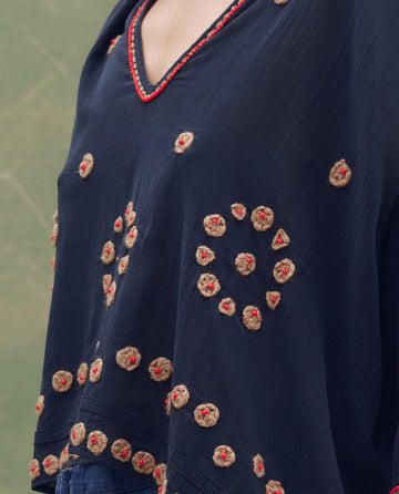 A person wears THE GREAT. SU25's The Beaded Vintage Sailor Top in navy, featuring red trim and circular tan and red beaded embroidery.