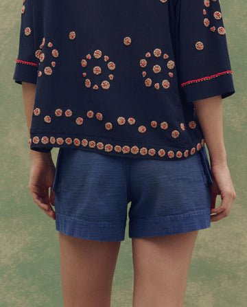 Person stands with their back to the camera, wearing a navy blue top with circular embroidery and THE GREAT. SU25 The Cargo Fatigue Short in Vintage Blue.
