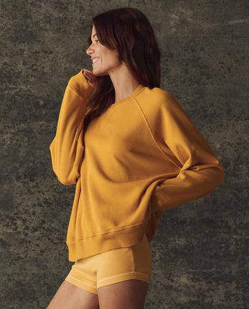 A woman stands sideways, smiling, in THE GREAT. The Fleece Slouch Sweatshirt Solid -- Bright Yellow (H25 BRIGHT FLEECEBACK), paired with matching shorts against a textured gray background.
