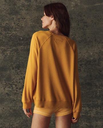 A person with long hair faces away, wearing THE GREAT.'s Bright Yellow Fleece Slouch Sweatshirt (H25 BRIGHT FLEECEBACK) and matching shorts made from soft French Terry, set against a textured dark background.