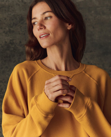 A woman in THE GREAT.’s The Fleece Slouch Sweatshirt in Bright Yellow (H25 BRIGHT FLEECEBACK) stands against a dark background, looking to the side with hands clasped.