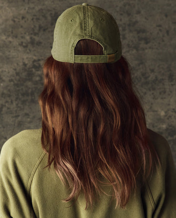 A person with long brown hair is seen from behind wearing THE GREAT. H25 BRIGHT FLEECEBACK Classic Baseball Hat in Army, paired with a matching sweatshirt, against a textured gray backdrop.