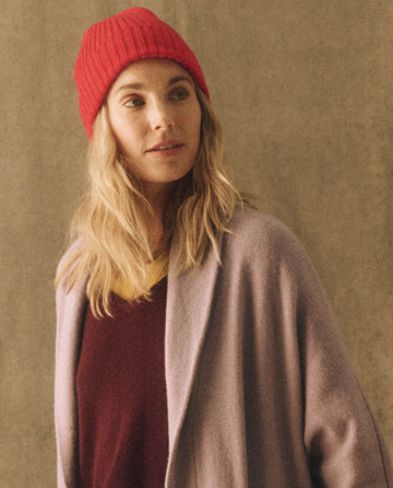 A person with long blonde hair wearing THE GREAT. HOL 22 D2 CASHMERE The Cashmere Hat in Bright Red, a maroon top, and a grey cardigan, looking slightly to the side.
