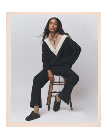 A woman sits on a wooden stool wearing a black outfit with a beige and white V-neck collar, tan socks, and black slippers against a plain, light background.